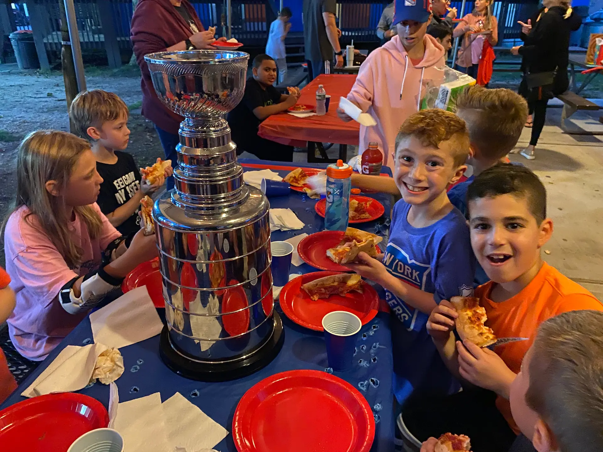 Children eat pizza at a table with a large trophy in the center, surrounded by red plates and drinks at an outdoor gathering.