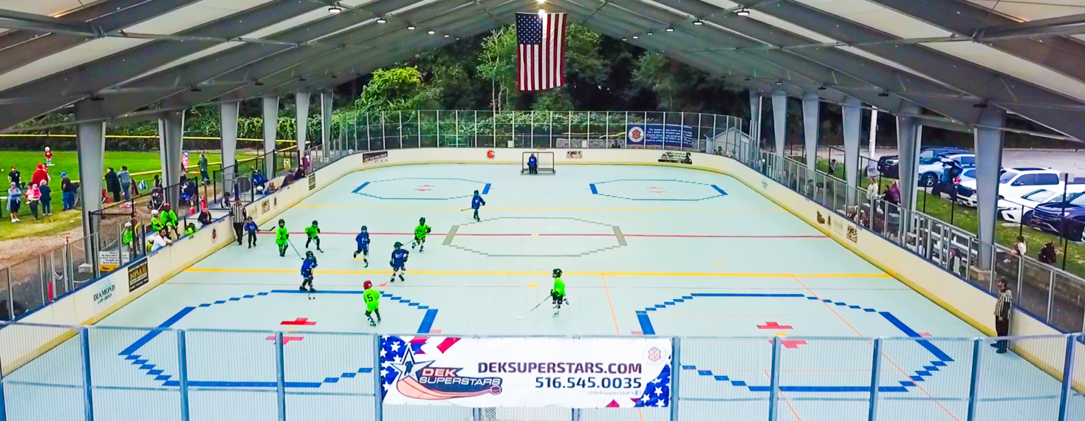 A youth dek hockey game is played under a covered outdoor rink, with teams in green and blue uniforms; an American flag hangs at the center above the rink.