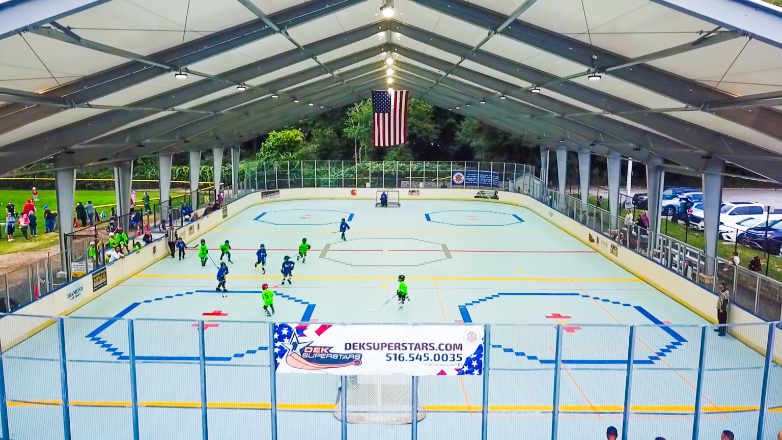 An outdoor covered roller hockey rink with children playing a game, spectators on the left, and an American flag hanging above the rink.